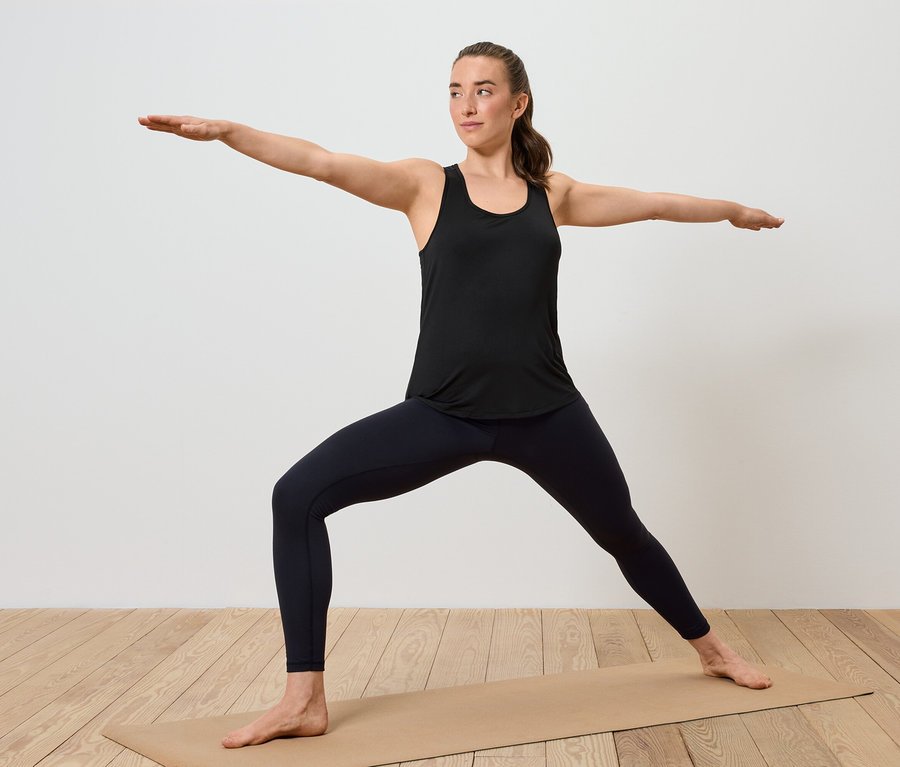 Frau in schwarzem Tanktop und Leggings steht in Krieger-II-Pose auf einer Yogamatte.