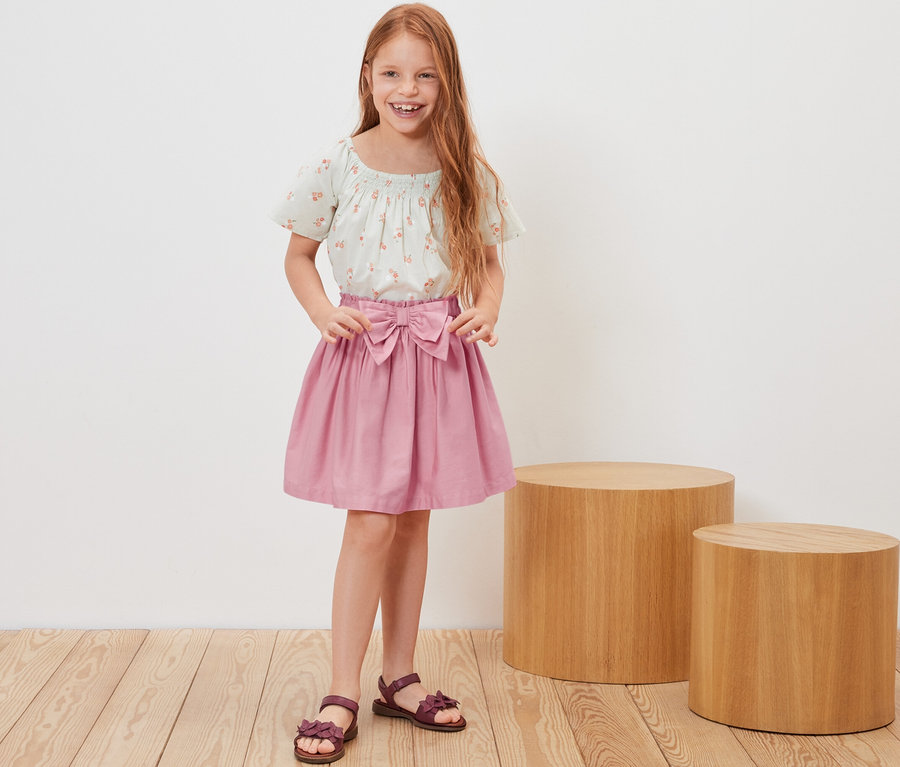 Lächelndes rothaariges Mädchen posiert in Kinder-Web-Rock und geblümtem Top.