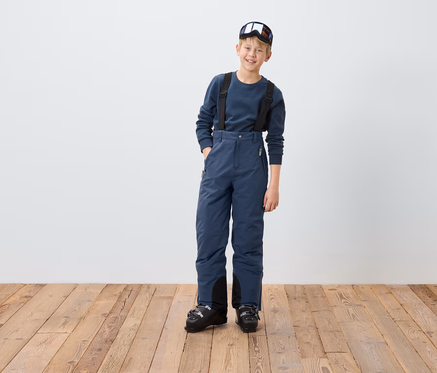 Ein Junge in blauer Skihose mit Hosenträgern und Skischuhen, mit einer Skibrille auf dem Kopf.