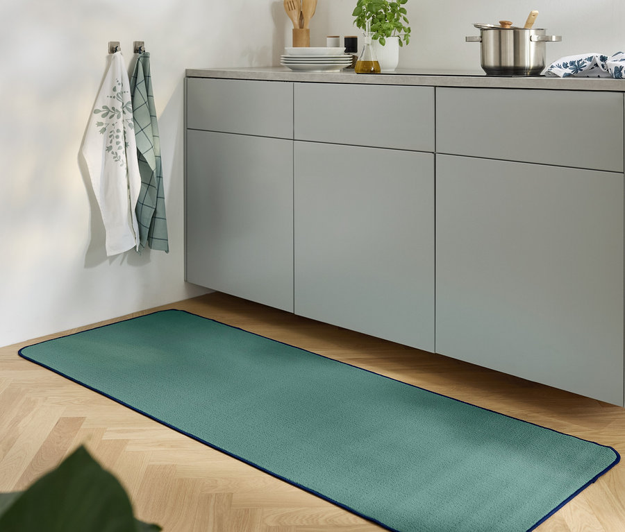 Ein grüner Teppich liegt auf einem Holzboden in einer Küche mit einem grauen Schrank.