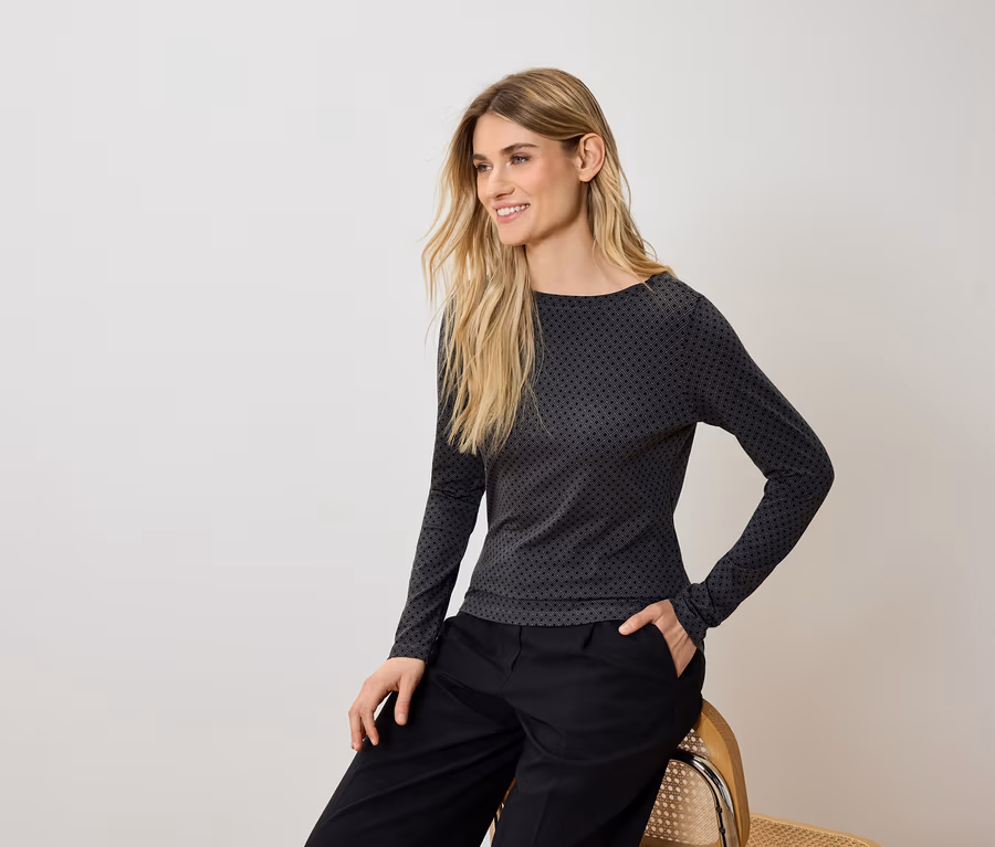 Blonde Frau sitzt auf einem Stuhl, trägt eine schwarze Hose und ein grau-schwarzes Langarmshirt.