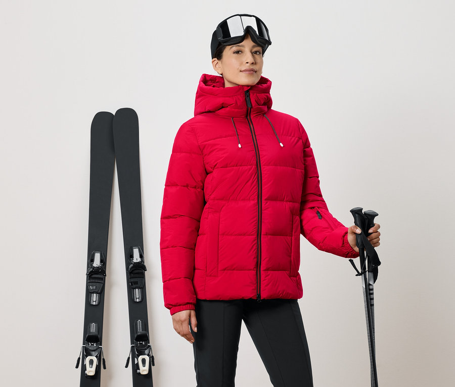 Frau in roter Skijacke hält Skistöcke, daneben Skier und eine Skibrille auf dem Kopf.