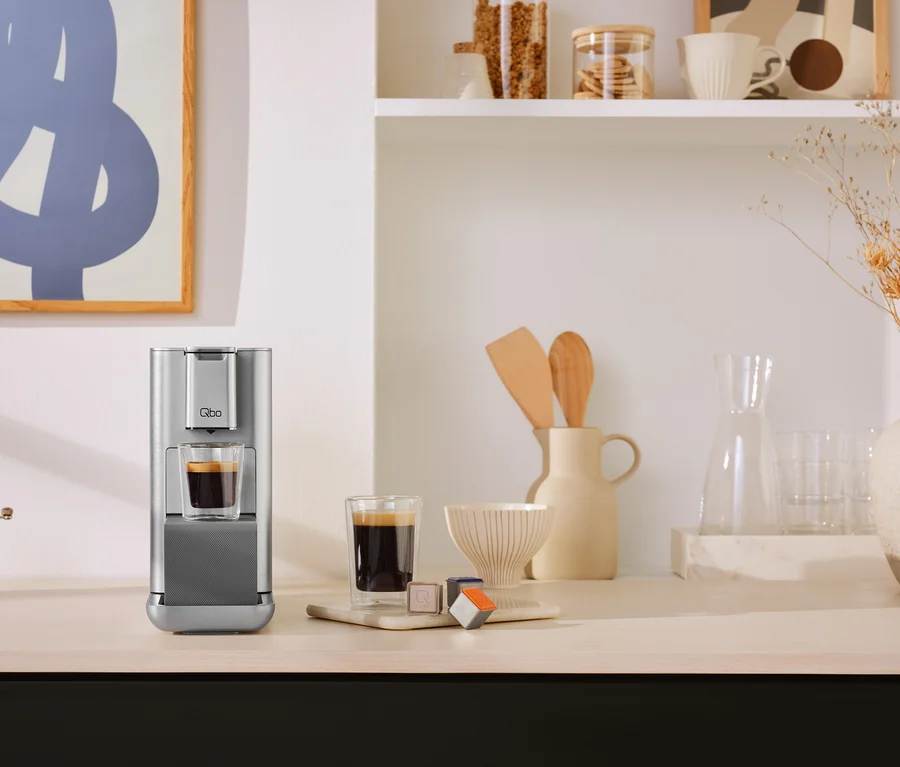 Silberne Kaffeemaschine auf einer Küchenarbeitsplatte, die Kaffee in ein Glas zubereitet. Daneben stehen ein weiteres Glas Kaffee und Kaffeekapseln. Im Hintergrund sind Regale mit Küchenutensilien und Dekoration zu sehen.