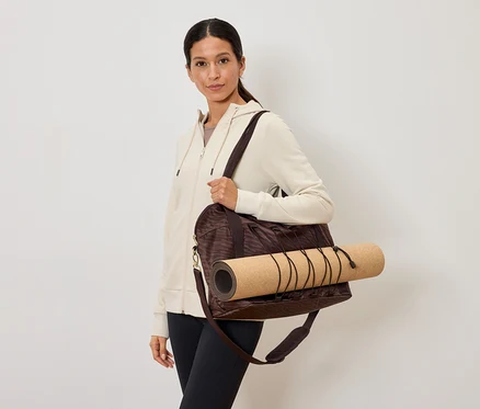 Frau trägt eine braune Sporttasche mit einer Yogamatte über der Schulter, dazu eine cremefarbene Jacke und schwarze Leggings.