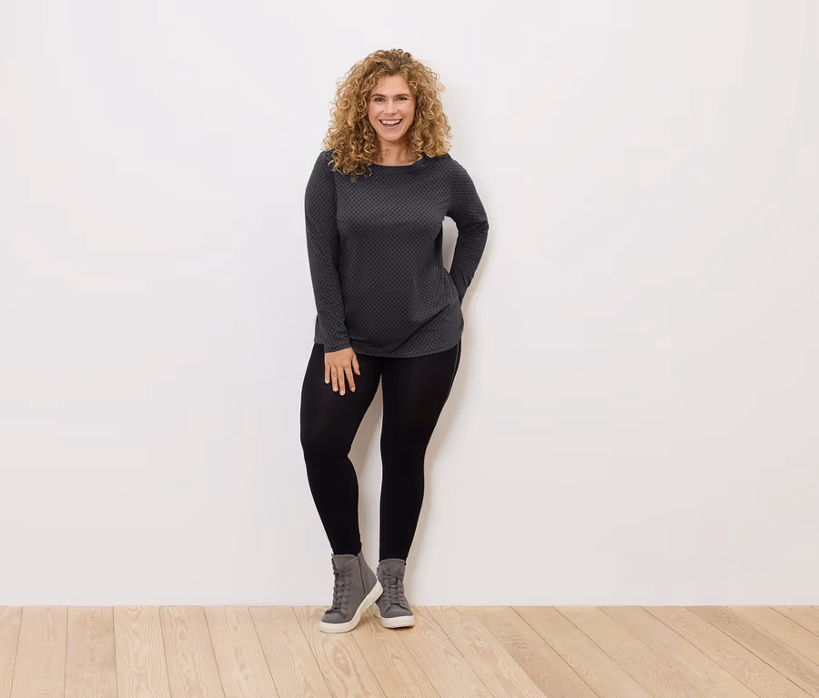 Frau mit lockigem Haar steht und lächelt, sie trägt schwarze Leggings, graue Schuhe und ein grau-schwarzes Shirt.