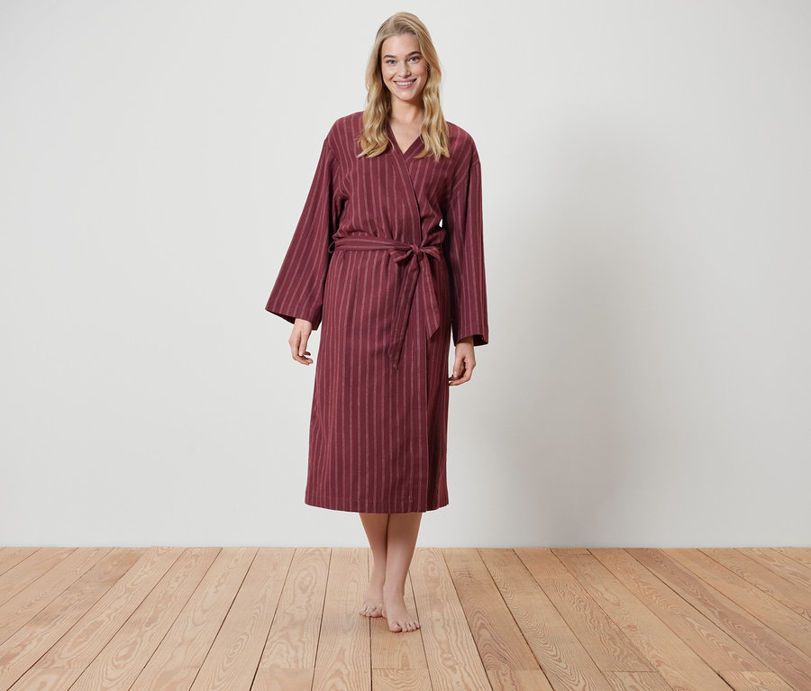 Eine blonde Frau steht auf einem Holzboden und trägt einen weinroten Bademantel mit Streifen.