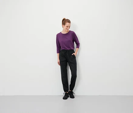 Frau mit braunen Haaren steht mit den Händen in den Taschen ihrer schwarzen Hose und trägt ein violettes T-Shirt.