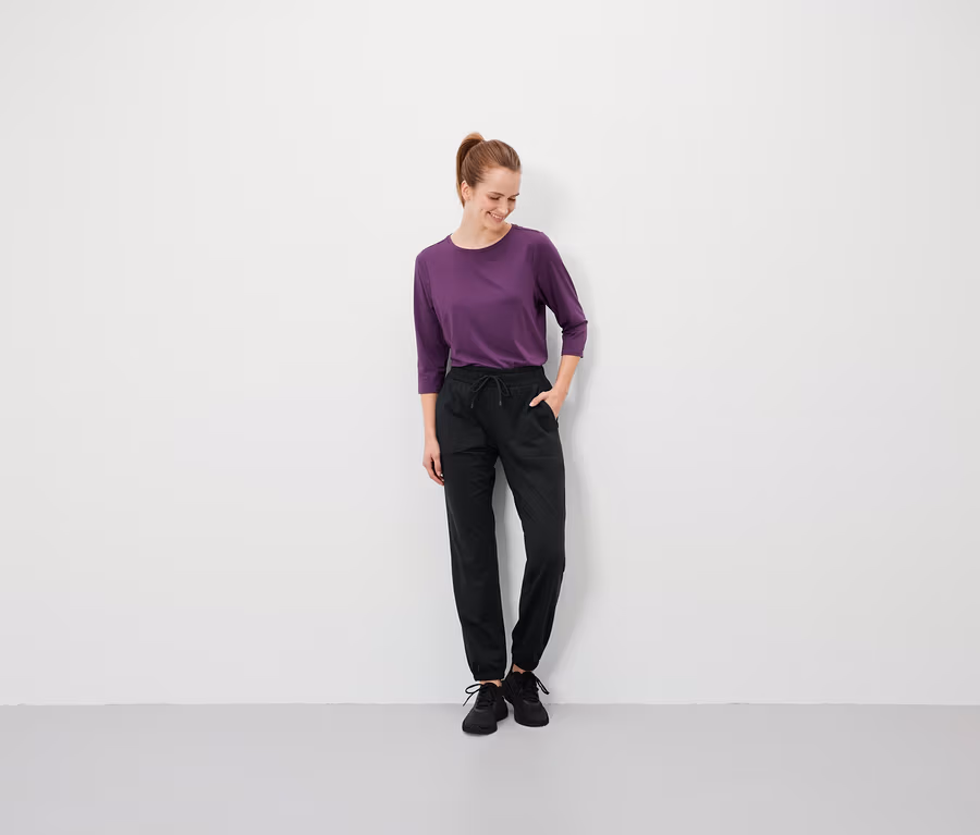 Frau mit braunen Haaren steht mit den Händen in den Taschen ihrer schwarzen Hose und trägt ein violettes T-Shirt.