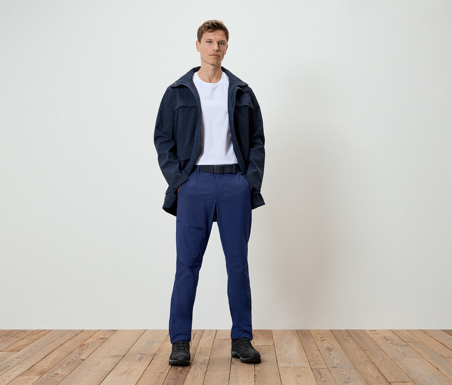 Ein Mann steht auf einem Holzboden. Er trägt eine dunkelblaue Jacke, ein weißes T-Shirt, blaue Hosen und schwarze Schuhe.