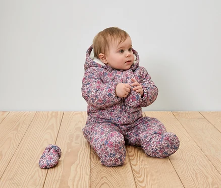 Ein Baby-Mädchen sitzt auf einem Holzboden in einem geblümten Winteranzug mit Kapuze und einem Schuh.