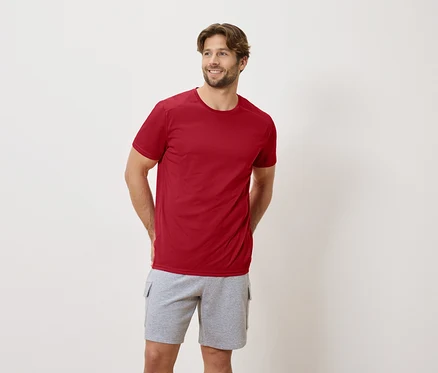 Ein Mann steht mit seinen Händen auf dem Rücken und trägt ein rotes T-Shirt und graue Shorts.