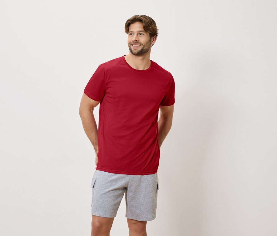 Ein Mann steht mit seinen Händen auf dem Rücken und trägt ein rotes T-Shirt und graue Shorts.