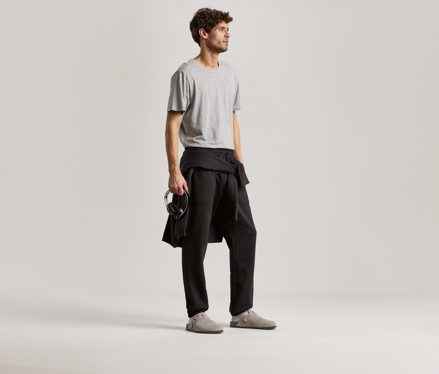 Mann trägt ein graues T-Shirt und eine schwarze Sweathose. Er hält Kopfhörer in der Hand.