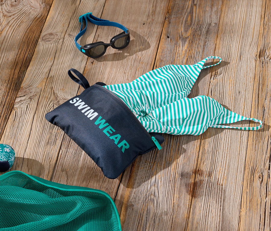 Aufnahme von Schwimmzubehör auf einer Holzoberfläche, darunter eine Tasche mit der Aufschrift "Swimwear", eine grün-weiß gestreifte Badehose, eine Schwimmbrille und ein grünes Netz.