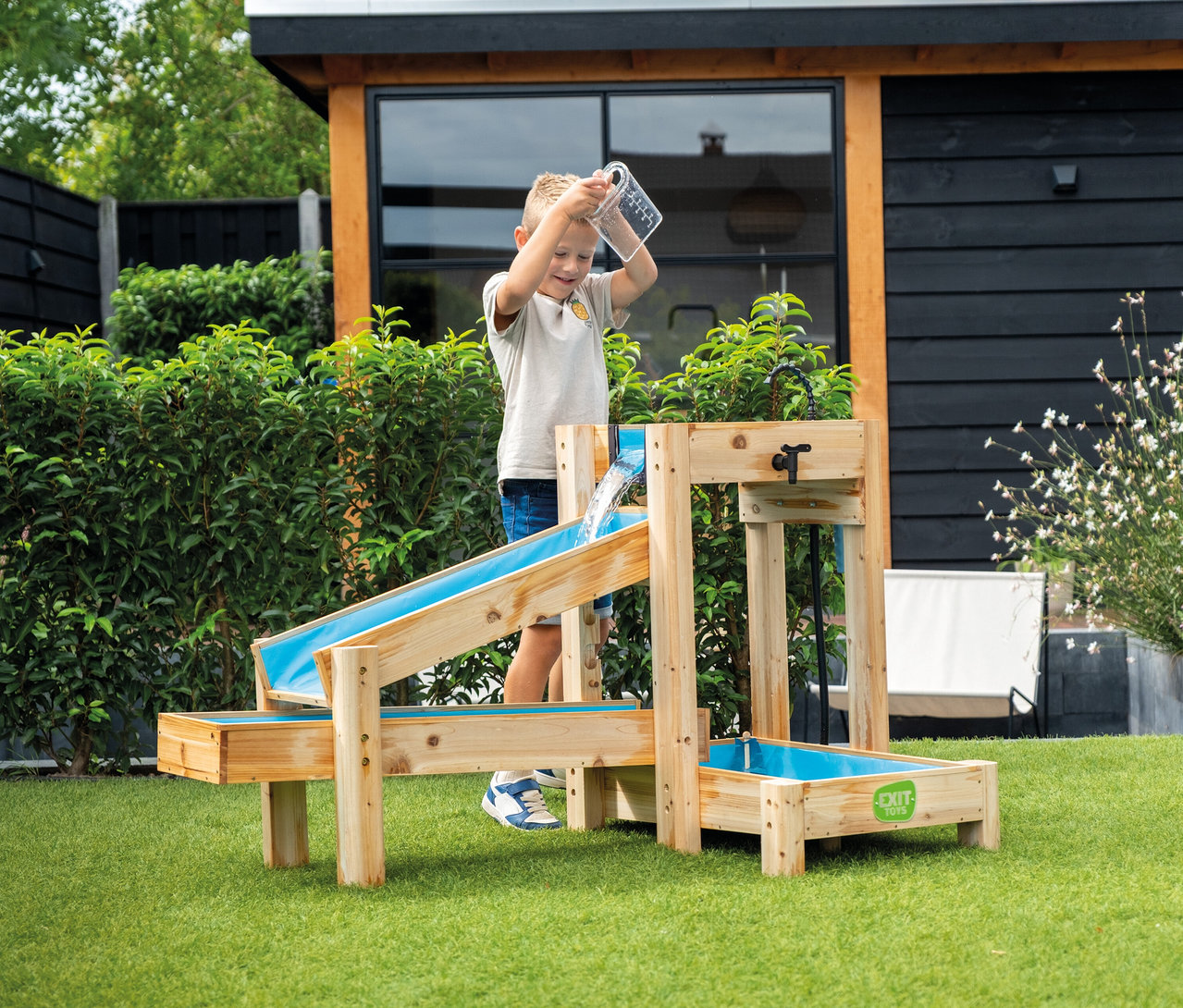 Junge gießt Wasser aus einem Messbecher in eine blaue Wasserrutsche auf einem Holzgestell im Garten.