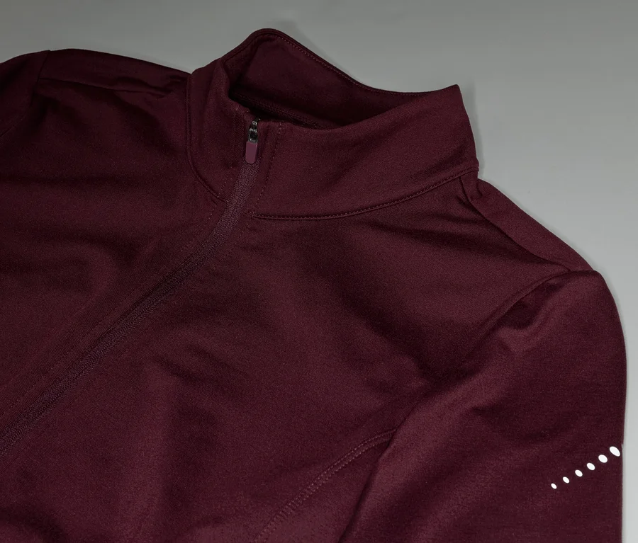 Nahaufnahme einer weinroten Trainingsjacke mit Reißverschluss und reflektierenden Punkten am Ärmel.