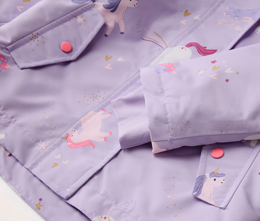 Nahaufnahme einer lila Jacke mit Einhornmuster, einer Tasche mit einem pinken Knopf und einem Ärmel.