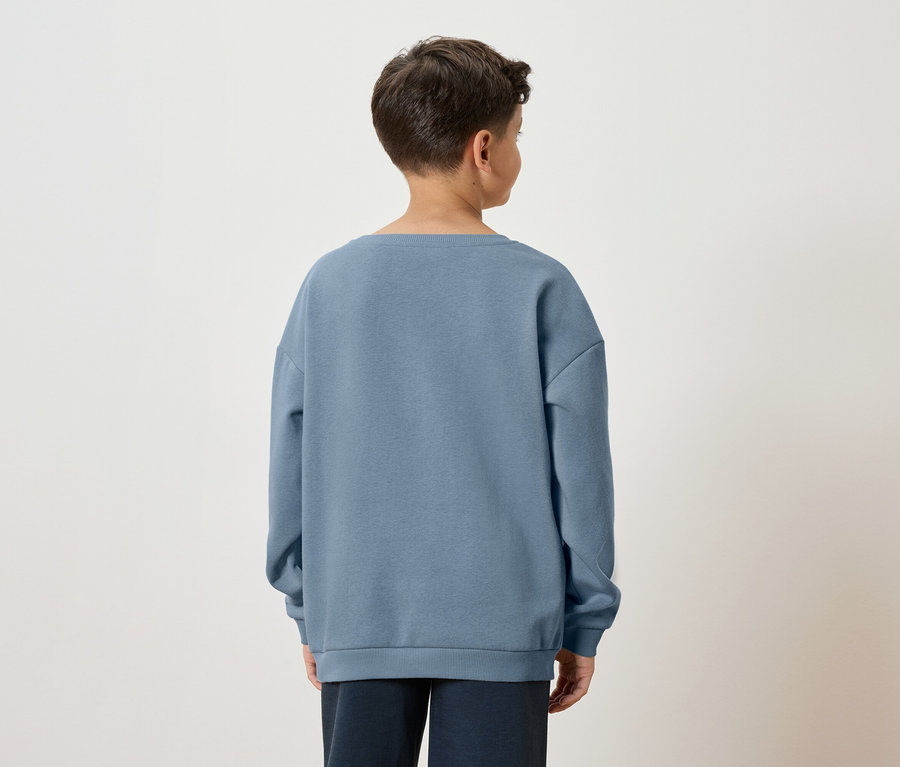 Rückansicht eines Jungen mit braunen Haaren, der einen blauen Pullover und eine dunkle Hose vor einem weißen Hintergrund trägt.