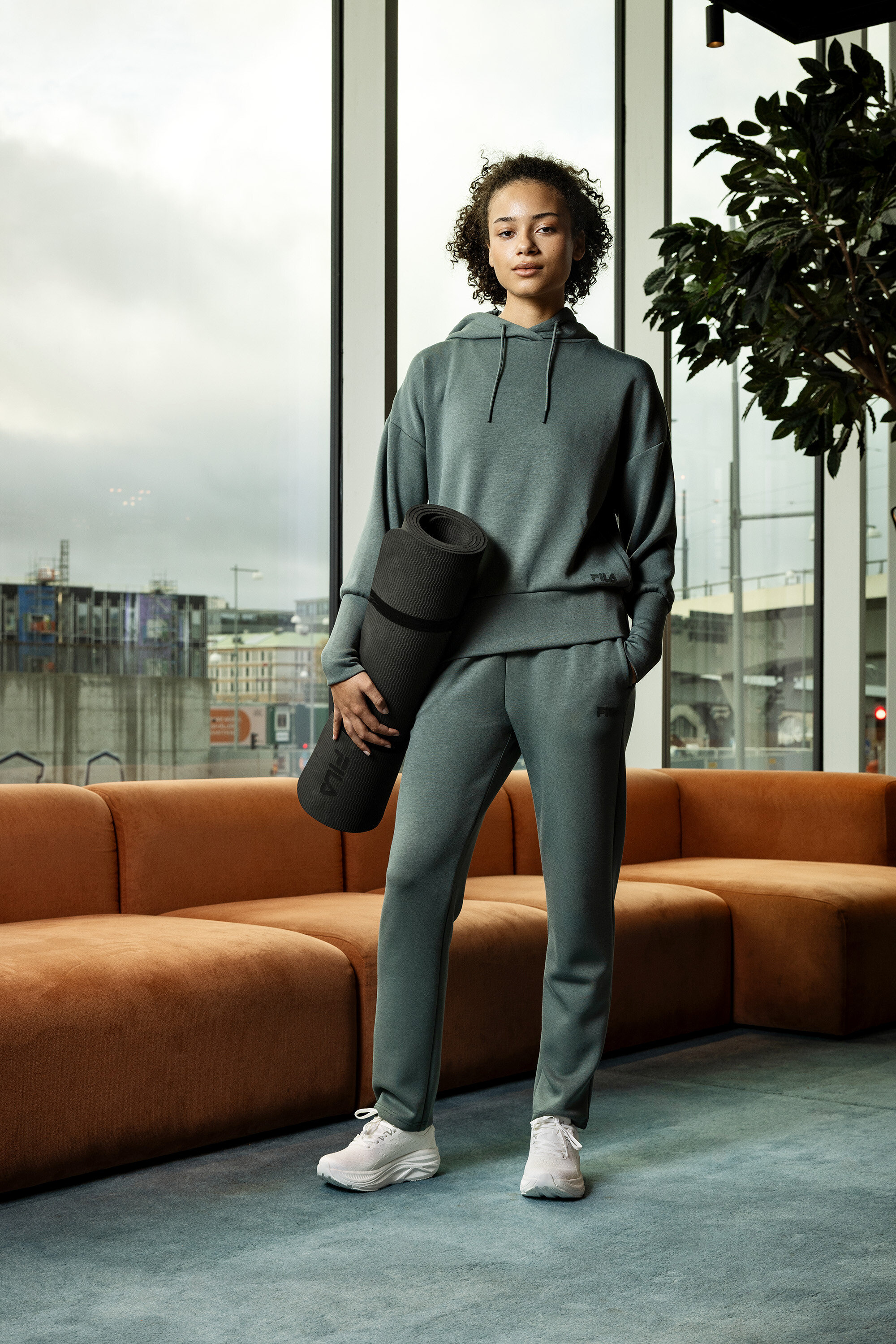 Junge Frau in grauer Trainingsbekleidung hält eine zusammengerollte Yogamatte vor einem Fenster.