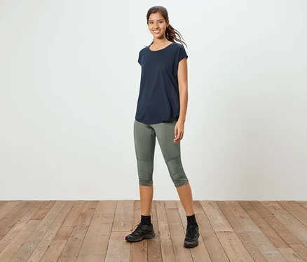 Eine Frau steht auf einem Holzboden, sie trägt ein marineblaues T-Shirt, eine olivgrüne 3/4-Hose und schwarze Turnschuhe.