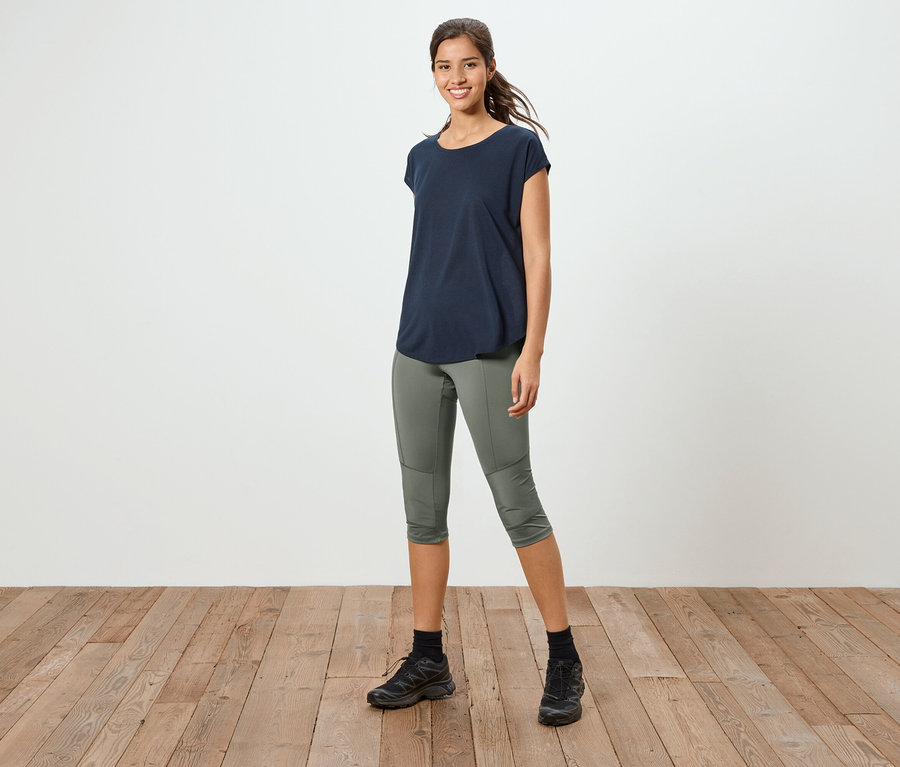 Eine Frau steht auf einem Holzboden, sie trägt ein marineblaues T-Shirt, eine olivgrüne 3/4-Hose und schwarze Turnschuhe.