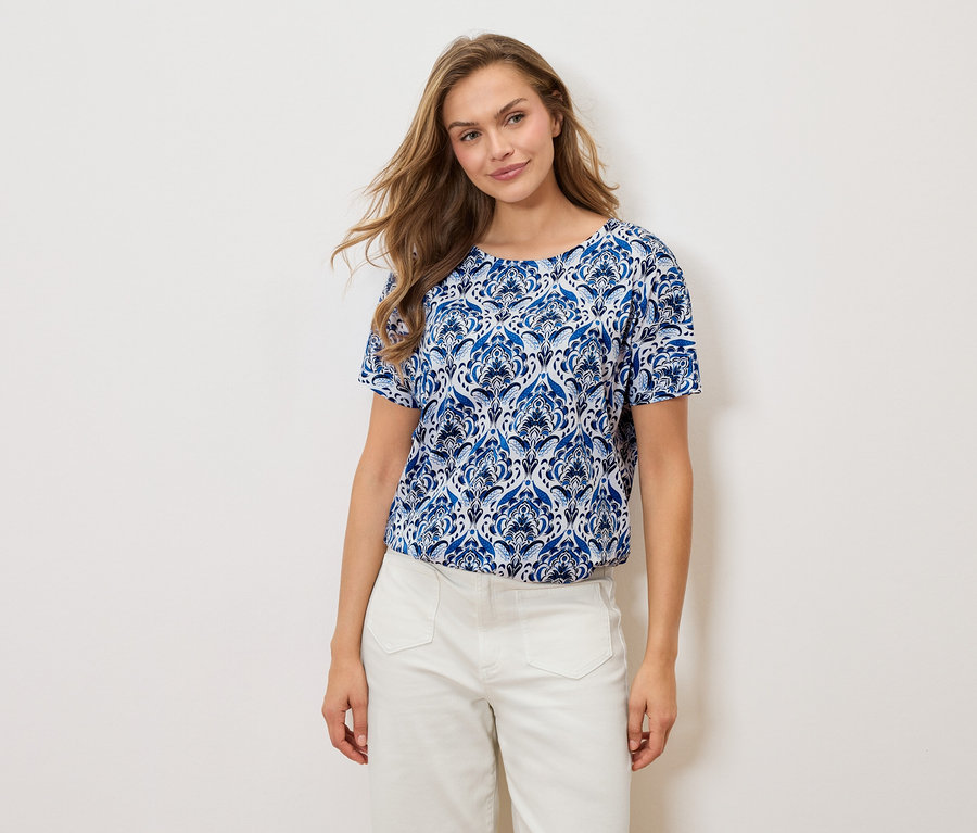 Frau trägt ein blau-weiß gemustertes T-Shirt und weiße Hosen vor einem weißen Hintergrund.