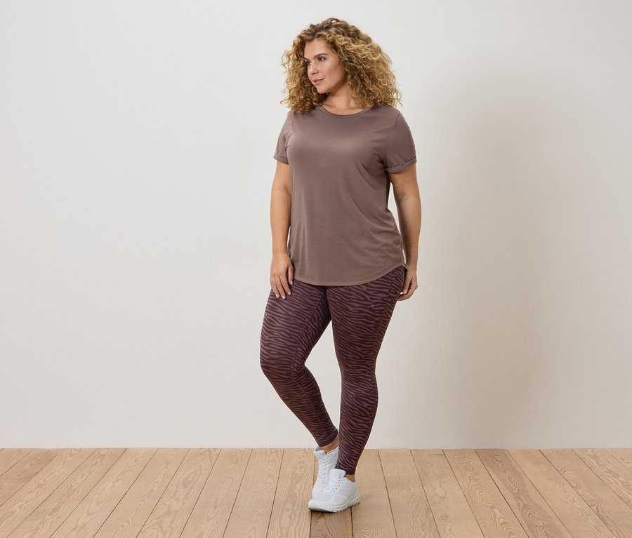 Frau mit lockigem Haar, braunem T-Shirt und gemusterten Leggings posiert vor einer Wand.