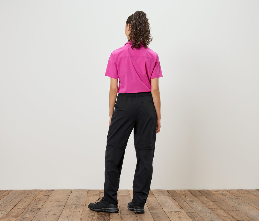 Rückansicht einer Frau mit lockigem Haar, die ein pinkes Hemd und schwarze, abzippbare Hosen über schwarzen Schuhen trägt.
