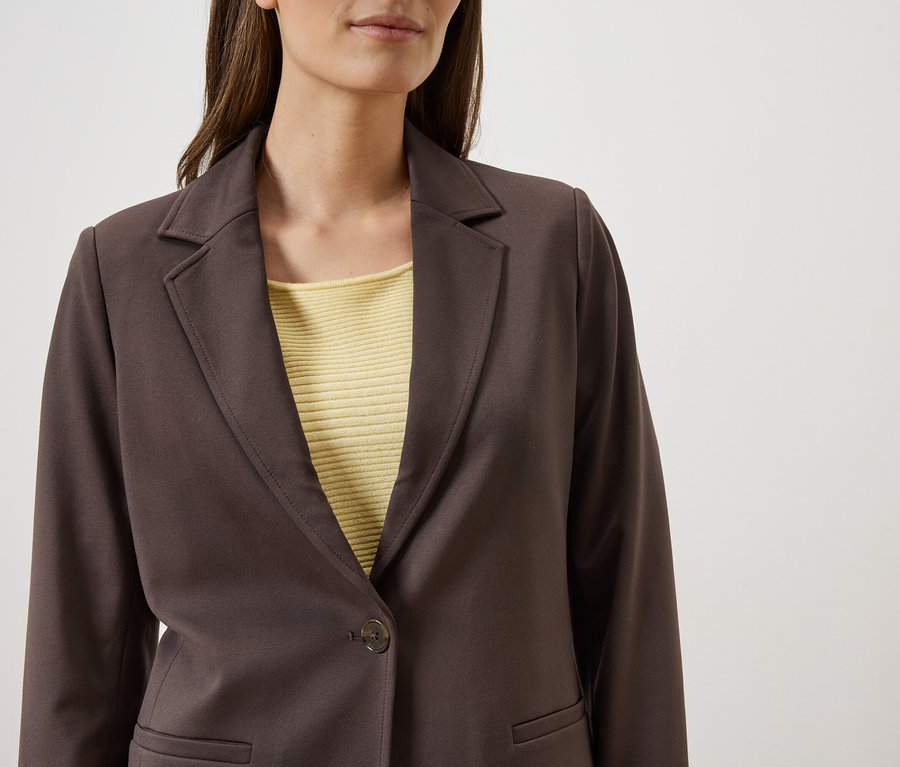Frau trägt braunen Blazer über einem gelben Oberteil vor einem weißen Hintergrund.