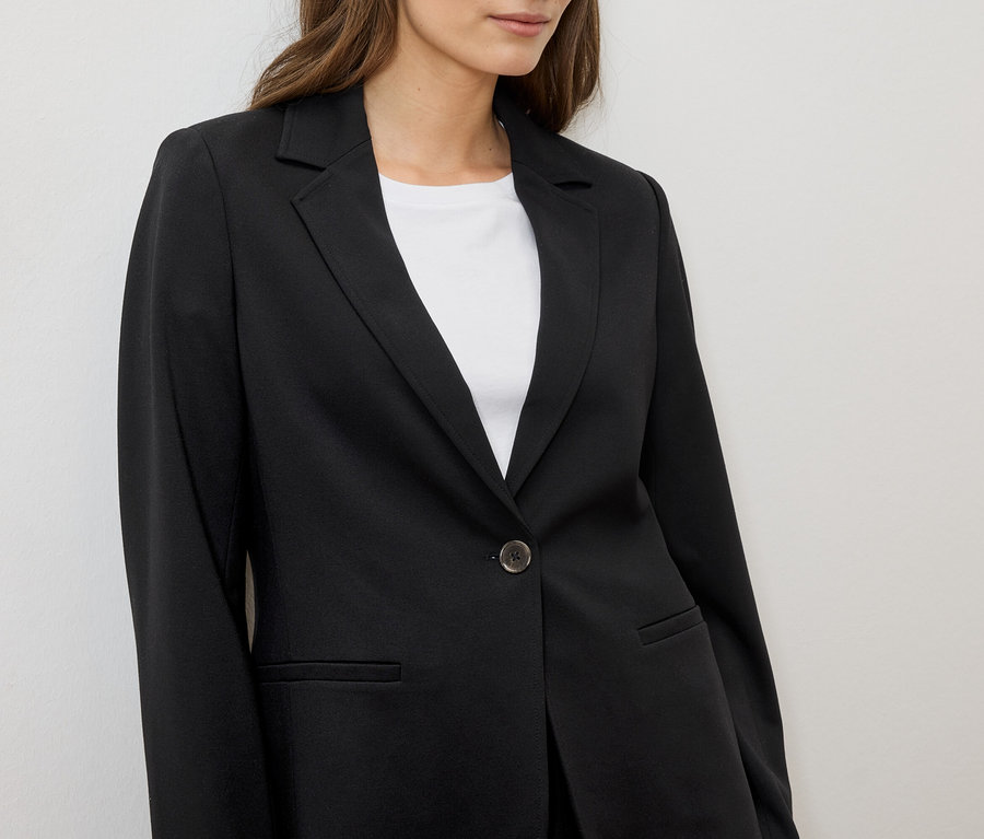 Frau trägt einen schwarzen Blazer über einem weißen T-Shirt vor einem weißen Hintergrund.