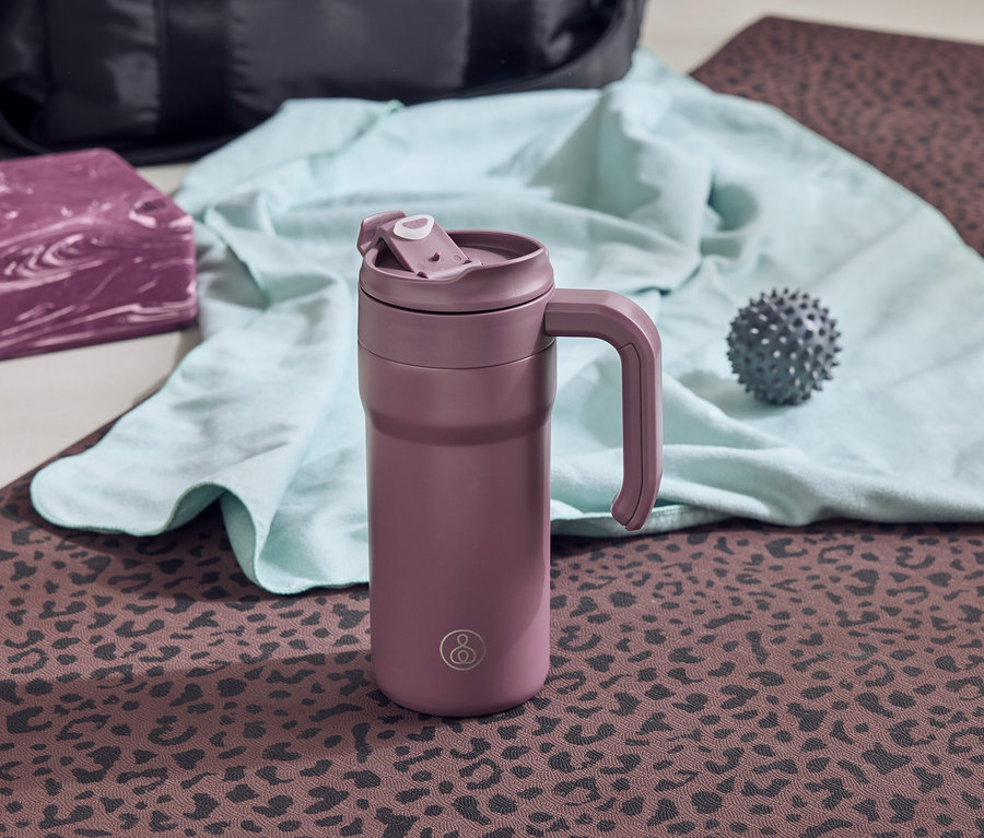Eine lila Thermoskanne mit Griff steht auf einer braunen Yogamatte mit Leopardenmuster. Daneben liegen ein grauer Igelball, ein türkisfarbenes Handtuch und ein lila Yogablock.
