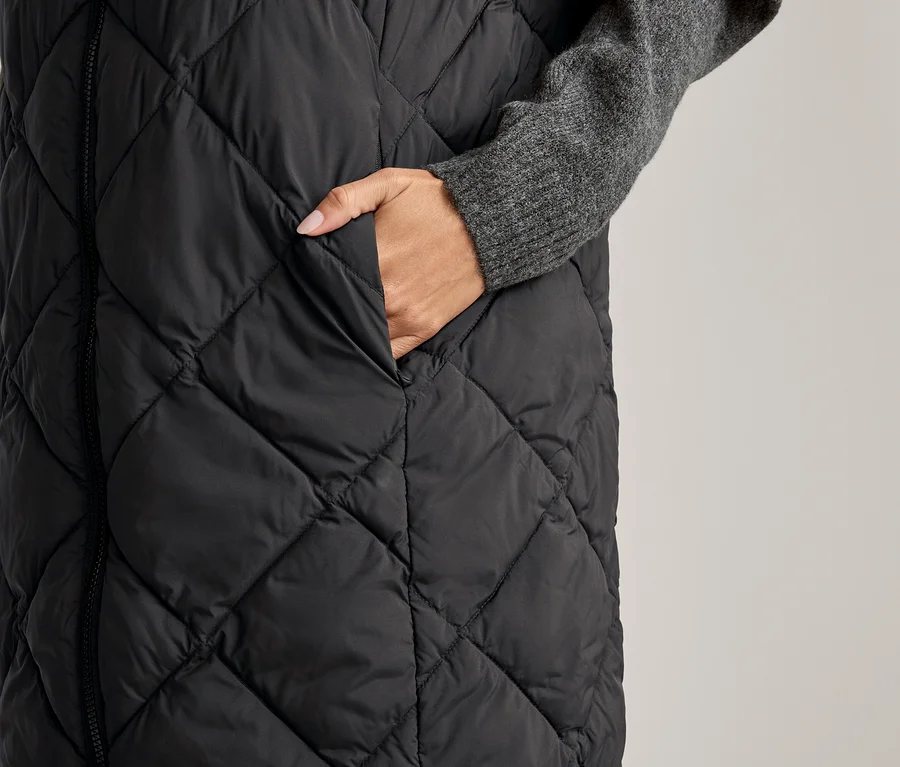 Frau steckt Hand in die Tasche einer langen, gesteppten, schwarzen Weste über einem grauen Pullover.