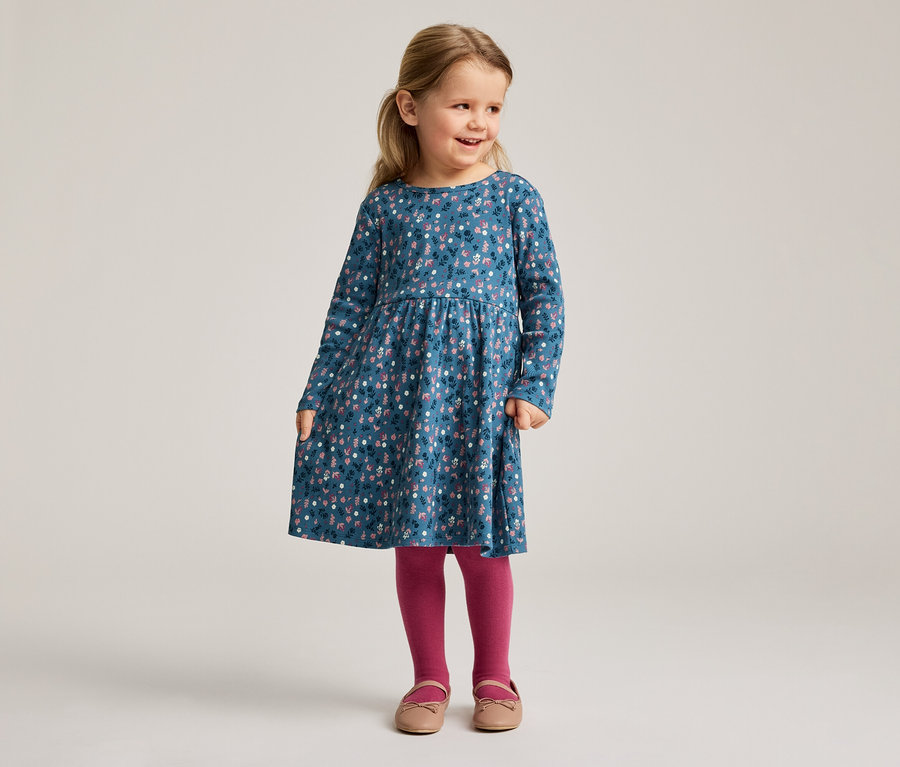 Ein kleines Mädchen mit blonden Haaren trägt ein blaues, langärmeliges Kleid mit Blumenmuster, dazu rosafarbene Strumpfhosen und Schuhe.