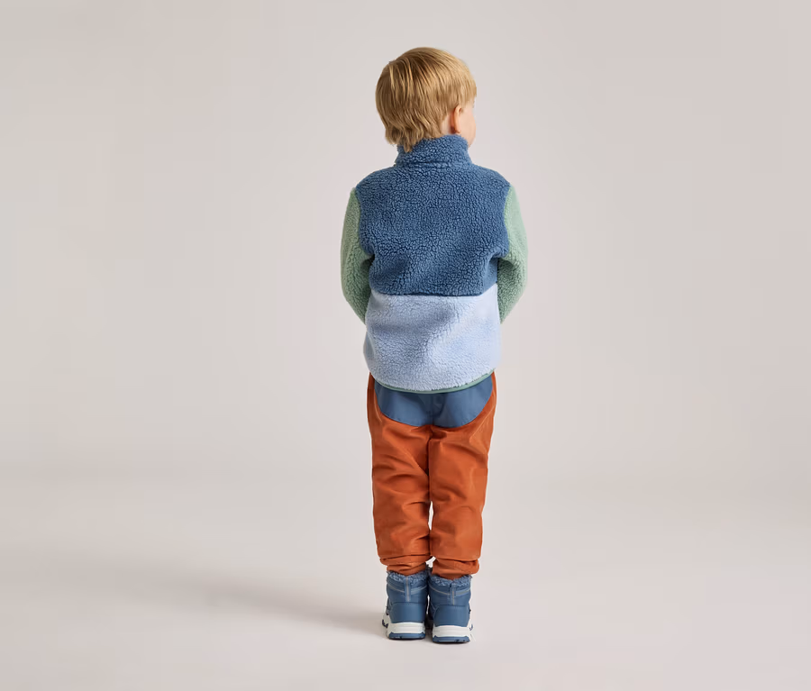 Rückansicht eines Kindes mit blonder Kurzhaarfrisur, das eine farblich abgesetzte Fleecejacke, orangefarbene Cordhosen und blaue Stiefel trägt.