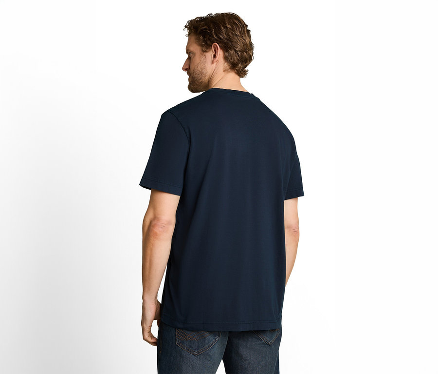 Ein Mann von hinten, der ein dunkelblaues T-Shirt und Jeans trägt und nach links blickt. Er hat kurzes braunes Haar und einen Bart.