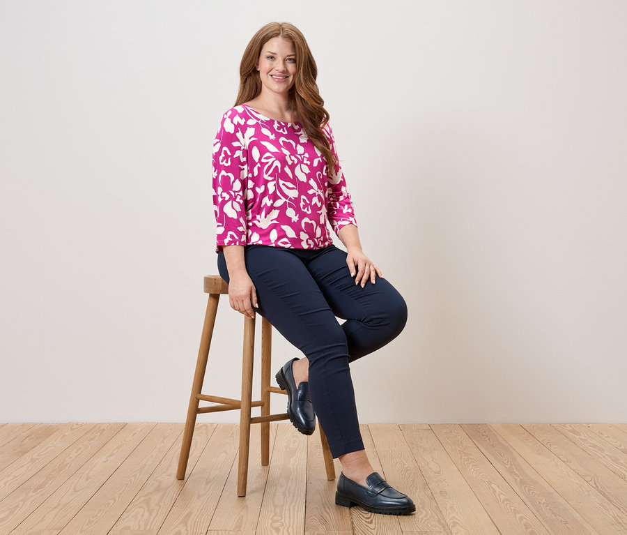 Frau sitzt auf einem Hocker, trägt eine pinkfarbene Bluse mit Blumenmuster, blaue Hosen und Lederschuhe.