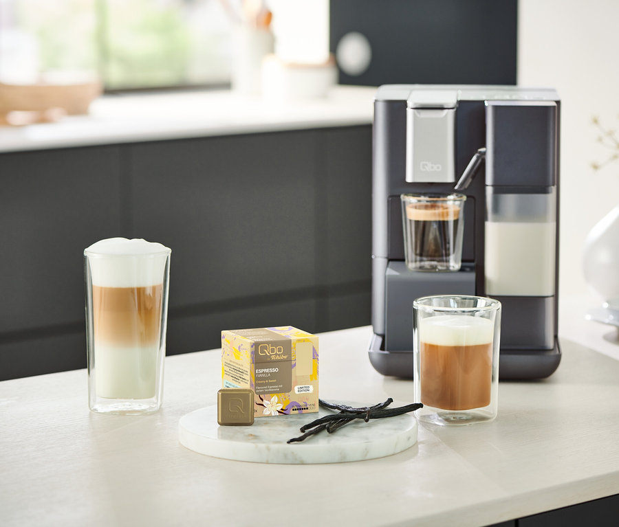 Ein Qbo MILK MASTER Gentle Grey steht neben zwei Gläsern Kaffee. Eine rote Qbo MILK MASTER ist im Hintergrund sichtbar.