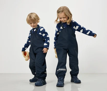 Zwei Kinder in marineblauen Overalls und blau-weißen Wolkenpullovern stehen auf einem weißen Hintergrund.