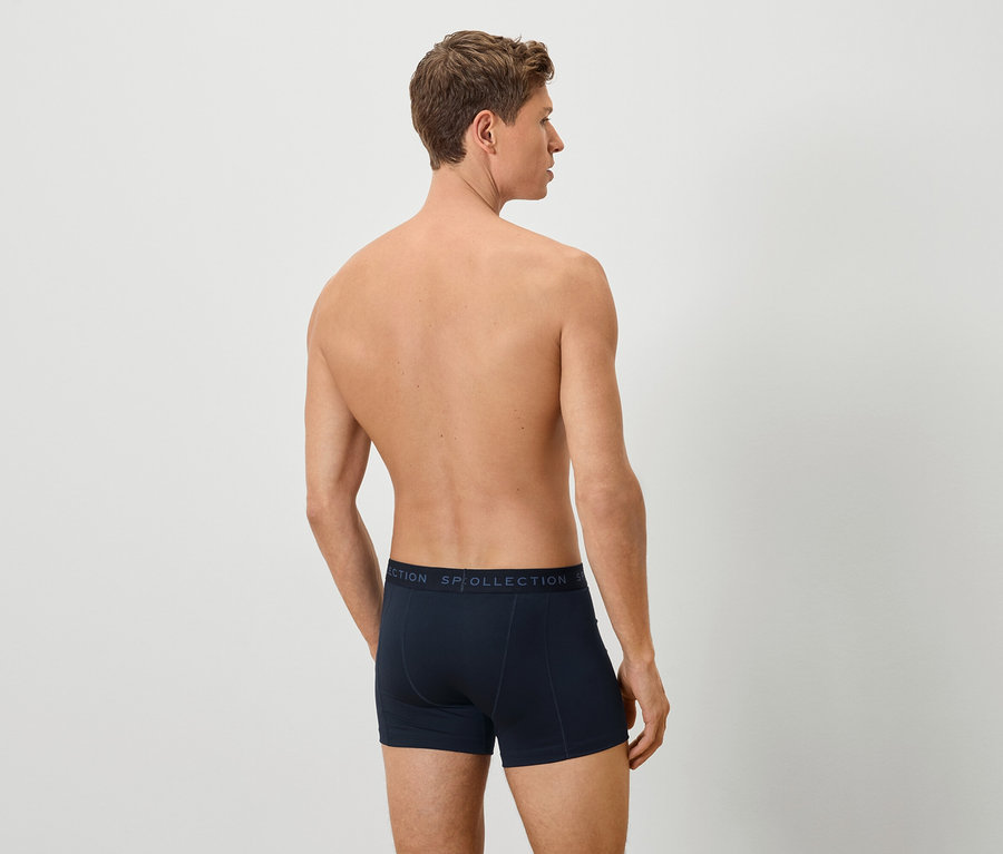 Rückansicht eines Mannes mit marineblauen Boxershorts vor einem neutralen Hintergrund.
