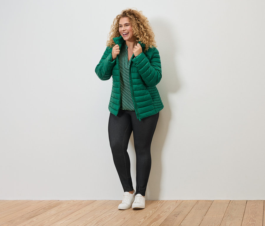 Frau mit lockigen Haaren, grüner Steppjacke, gestreifter Bluse, dunklen Leggings und weißen Turnschuhen.