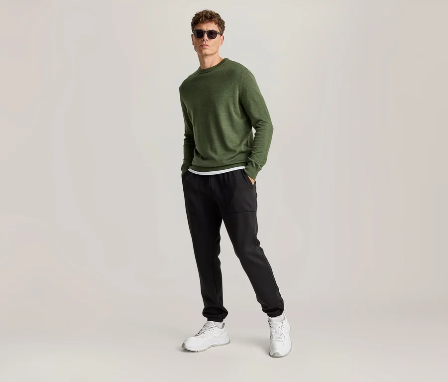 Mann trägt grünes Langarmshirt, schwarze Hose, weiße Sneaker und Sonnenbrille, steht mit Händen in den Hosentaschen.