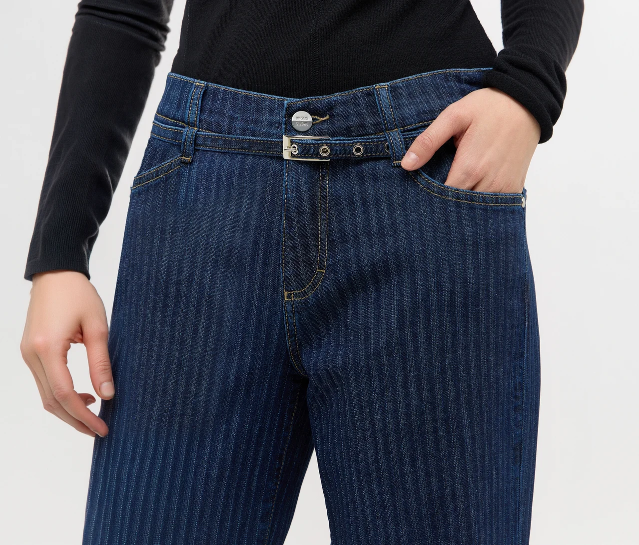 Person in dunkelblauer Jeans mit dezenten Längsstreifen und schwarzem Langarmshirt. Eine Hand steckt in der Hosentasche. Die Jeans hat einen Gürtel mit silberner Schnalle und einen silbernen Knopf.