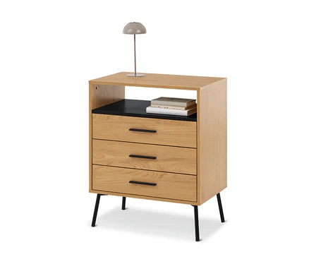 Nachttisch aus hellem Holz mit drei Schubladen und einem offenen Fach, darauf eine graue Lampe und Bücher.