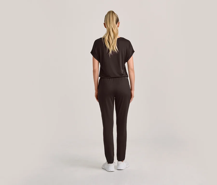 Frau von hinten in einem dunklen, kurzärmeligen Overall mit Gummizug in der Taille, blondes Haar im Pferdeschwanz und weiße Sneaker.