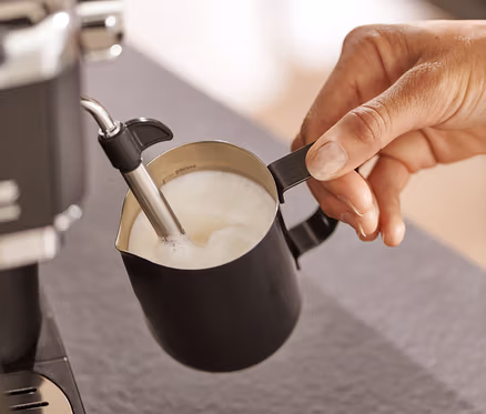 Person schäumt Milch in einem schwarzen Milchkännchen mit einer Kaffeemaschine auf.