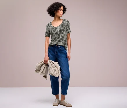 Eine Frau mit kurzen, lockigen dunklen Haaren trägt ein schwarz-weiß gemustertes Street One T-Shirt, blaue Jeans und helle Sneaker. Sie hält eine hellbeige Jacke in ihrer linken Hand und blickt nach rechts.