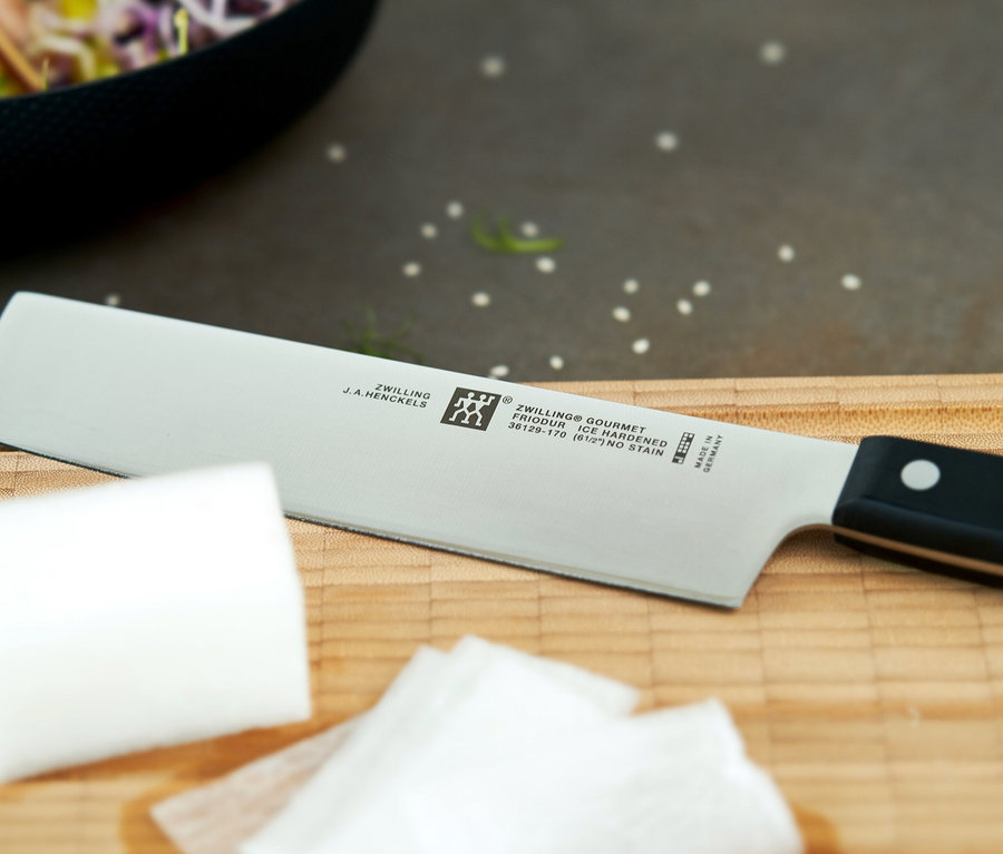 Ein Zwilling Gourmet Küchenmesser mit schwarzem Griff und silberner Klinge liegt auf einem Holzschneidebrett. Daneben liegen weiße Lebensmittelstücke, möglicherweise Käse oder Tofu. Im unscharfen Hintergrund ist eine schwarze Schüssel mit Salat zu sehen.