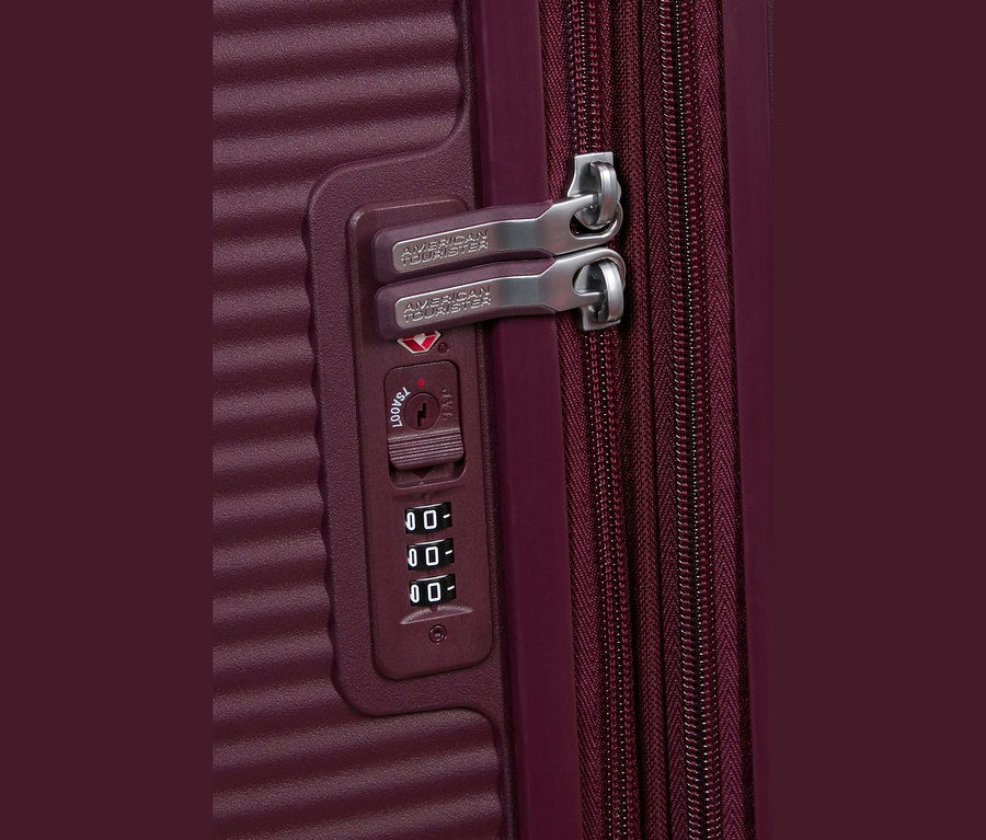 Nahaufnahme eines bordeauxroten American Tourister Koffers mit integriertem Zahlenschloss. Zwei silberne Reißverschlusszipper, jeweils mit „AMERICAN TOURISTER“ graviert, stecken im Schloss. Darunter befinden sich ein TSA007 Schlüsselloch und ein dreistelliges Zahlenrad, das auf „000“ eingestellt ist.