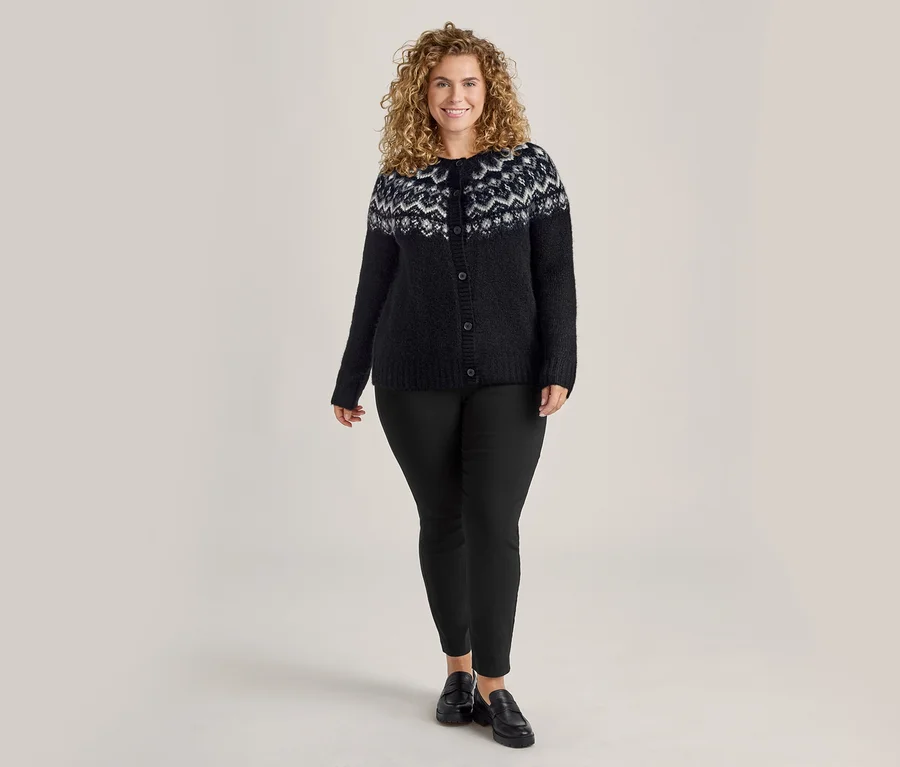 Frau mit lockigem Haar, die eine schwarze Strickjacke mit weißem Muster und eine schwarze Stretchhose trägt.