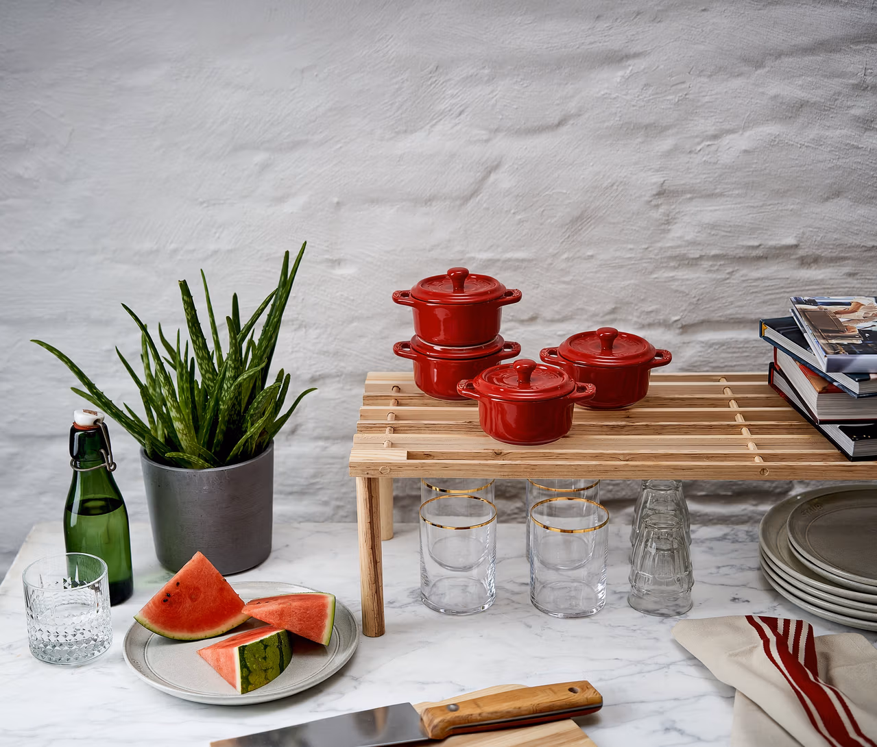 Drei kirschrote STAUB Keramik-Cocottes auf einem Holzregal, daneben Wassermelonenscheiben, eine Pflanze und Gläser auf einer Marmorplatte.